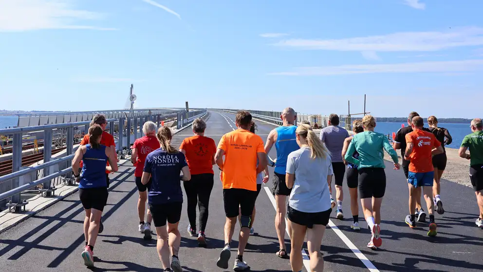 En gruppe mennesker iført sportstøj løber over en bro i solskinsvejr.